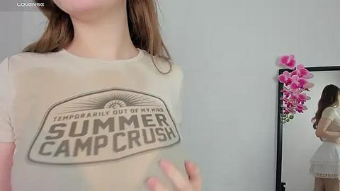 Media: Video of a woman with long brown hair, wearing a see-through white T-shirt with \"Summer Camp Crush\" text, standing in front of a mirror reflecting her back, with pink orchids in the background.