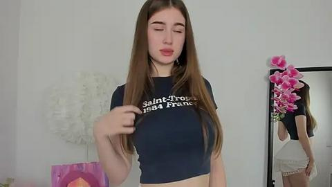 Media: Video of a young woman with long brown hair, wearing a black crop top with white text, standing in a minimalist room with pink flowers and a mirror.