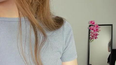 Media: Video of a light-skinned woman with shoulder-length, straight brown hair in a light blue, ribbed t-shirt. Background features a mirror with pink flowers and a partially visible person in a black shirt.