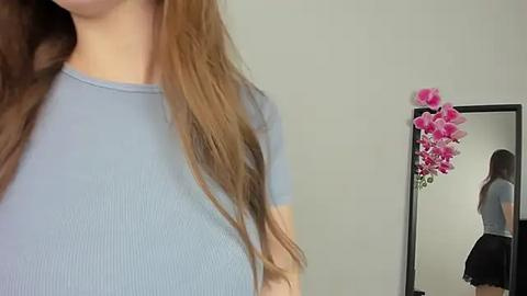 Media: Video of a young woman with long, wavy, light brown hair, wearing a tight, light blue ribbed top, standing in front of a mirror reflecting her backside in a black skirt. A pink orchid is placed in the mirror.