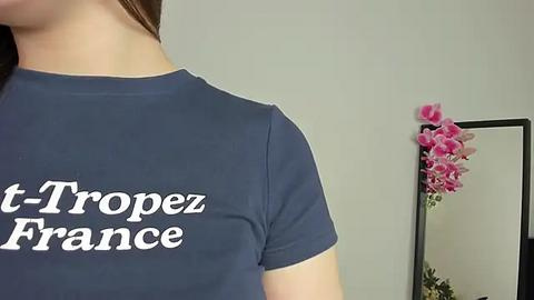 Media: Video of a woman with fair skin wearing a navy blue t-shirt with white text \"t-Trop\u00e9zance\" on the chest, partially seen. Background features a mirror with pink orchids and a plain white wall.