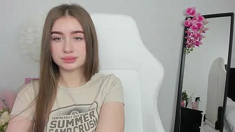 Media: Video of a young Caucasian woman with straight brown hair, light skin, wearing a beige T-shirt with a vintage design, sitting on a white gaming chair in a minimalist room with a pink flower arrangement and a mirror.