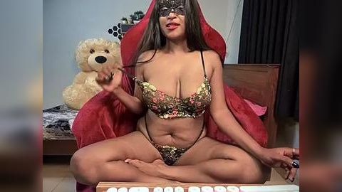 Media: Video of a light-skinned woman with long dark hair, wearing a black lace mask, floral lingerie, and a red blanket, sitting cross-legged on a wooden floor, playing chess, with a plush bear in the background.