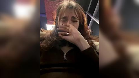 Media: Video of a woman with shoulder-length, light brown hair, crying, holding a smartphone to her face, wearing a dark coat, in a dimly lit room with blurred people in the background.