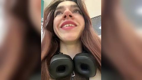 Media: A close-up video of a smiling young woman with long brown hair, wearing black headphones, taken from a low angle, with blurred human hands in the foreground.