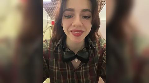 Media: Video of a young woman with light skin and brown hair, smiling. She wears a plaid shirt, black bow tie, and black headphones, sitting in an office with beige walls and a ceiling grid.