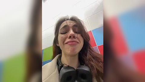 Media: Video of a young woman with long brown hair, closed eyes, and a slight smile, wearing black headphones, in a tiled bathroom with colorful tiles.