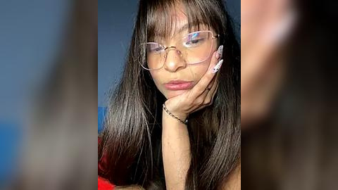 Media: Video of a young woman with long, straight black hair and fair skin, wearing round glasses and a red dress, resting her chin on her hand, with blurred background.