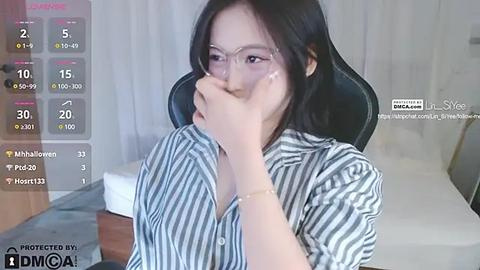 Media: A candid video of an Asian woman with shoulder-length black hair and glasses, wearing a striped shirt, covering her mouth, and seated on a black chair in a modern room.