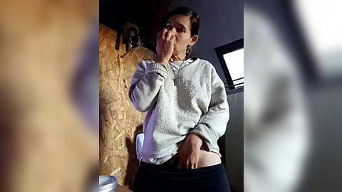 Media: A video shows a woman with dark hair, wearing a white sweater and black pants, covering her mouth in a bathroom with a rustic wall and a sink.