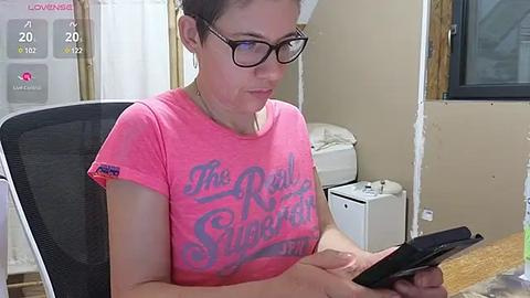 Media: Video of a fair-skinned woman with glasses, pink t-shirt, and short hair, sitting at a desk, holding a tablet. Background includes beige walls, a window, and a white printer.