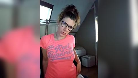 Media: Video of a young woman with glasses, wearing a pink t-shirt with a \"The Royal Tenenbaums\" quote, sitting in a dimly-lit room with a small white printer on the right.