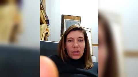 Media: A video of a woman with light skin and shoulder-length brown hair, wearing a black sweater, seated in a dimly lit room with framed pictures and a wooden chair in the background.