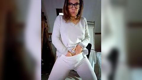 Media: Video of a woman in white pajamas, with glasses, standing in a dimly lit bedroom, holding a black object.