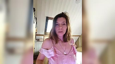 Media: A video of a Caucasian woman with shoulder-length brown hair, wearing a pink off-shoulder top and a necklace, seated indoors with a blurred background.