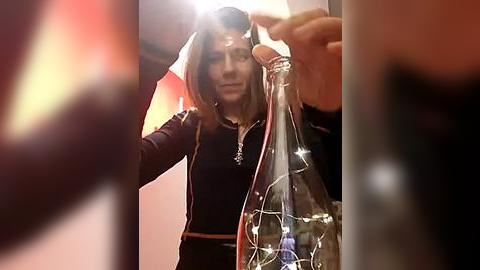 Media: Video of a woman with shoulder-length brown hair, wearing a black jacket, holding a clear glass bottle with twinkling lights inside, against a warm-toned indoor background.