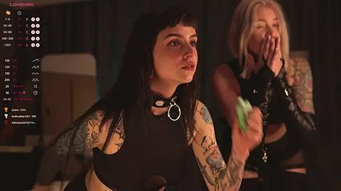 Media: Video: A tattooed woman with black hair, wearing a black crop top, sits indoors. A blonde woman in a black leather jacket stands behind her, covering her face.