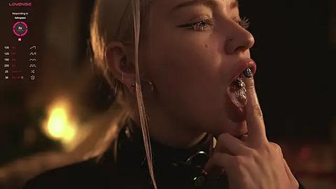 Media: Video of a young woman with pale skin, blonde hair, and pierced nose, in a dimly lit room, holding a finger to her mouth, with a muted background.