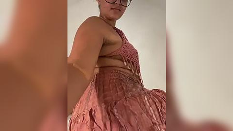 Media: Video of a plus-sized woman with medium skin tone, wearing a pink crocheted halter top and matching skirt, standing against a plain beige wall.