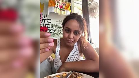 Media: A video of a young woman with medium brown skin and curly hair, wearing a white crochet top, eating at a restaurant. Background includes a blurry figure and a colorful banner.