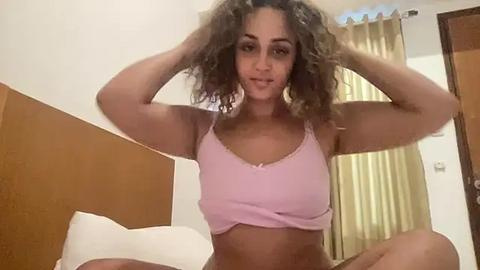 Media: Video of a light-skinned woman with curly hair, wearing a pink crop top, sitting on a bed, hands in her hair, in a beige room with a wooden headboard and curtains.