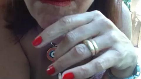 Media: Video of a woman with light skin, wearing red nail polish, touching her lips with her hand adorned with a ring, necklace, and a circular pendant.