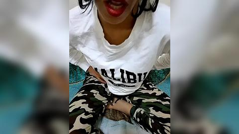 Media: A video showing a woman with medium skin tone, wearing a white T-shirt with \"SALIBI\" in black, and camouflage pants, sitting on a teal carpet.