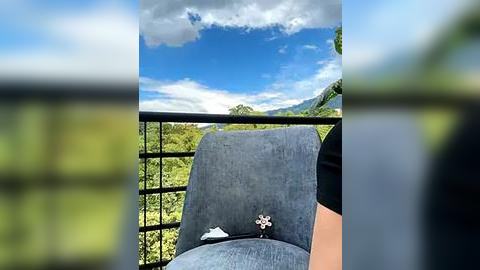 Media: Video of a woman sitting on a gray, textured chair on a balcony, with a mountainous landscape and a blue sky with clouds visible in the background.