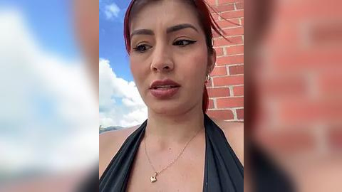 Media: Video of a Latina woman with fair skin, red hair, and dark eyebrows, wearing a black halter top, standing against a brick wall.