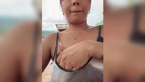 Media: Video of a woman with light skin, medium build, and medium-sized breasts wearing a gray tank top, with a small tattoo on her left arm, standing outdoors by a pool with greenery in the background.