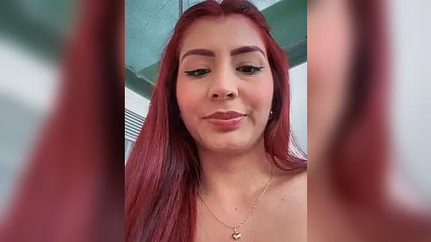 Media: Video of a young Latina woman with long, straight red hair, wearing black eyeliner, light makeup, and a gold necklace. She has a light complexion and is smiling.