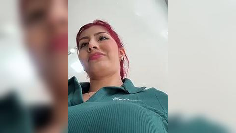 Media: Video of a confident, smiling woman with light skin and red hair, wearing a green ribbed polo shirt.