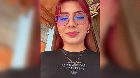 Media: Video of a woman with red hair and blue eyeshadow, wearing glasses and a black Creative Studio tee, indoors with a rustic background.