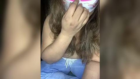 Media: A video of a young woman with long brown hair, wearing a white mask covering her face, holding it with her right hand. She is dressed in a light blue tank top and light blue pajama pants, seated on a bed.