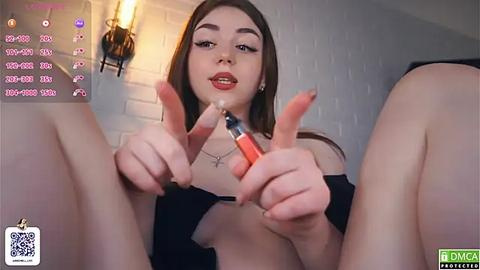 Media: A young woman with fair skin and brown hair, wearing a black top, spreads her legs, holds a red lipstick, and gives a thumbs-up gesture.