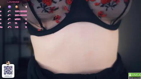 Media: A video of a woman lifting her sheer, floral-patterned top to reveal her midsection, showcasing a black bra and lower abdomen, taken in a dimly lit room.