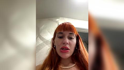 Media: Video of a fair-skinned woman with long, straight red hair, wearing a pearl necklace, looking down, in a dimly lit room with white walls.