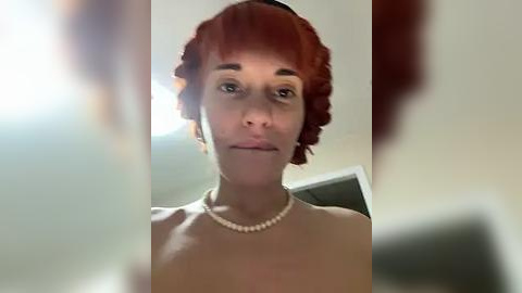 Media: A video of a topless woman with short, curly red hair, wearing a pearl necklace. She has fair skin, light makeup, and is indoors with a blurred background.