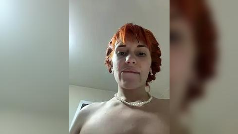 Media: A video of a fair-skinned person with short, curly, vibrant red hair, wearing a pearl necklace, smiling. The background is blurred, showing a white wall.