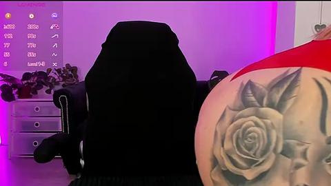 Media: Video of a black gaming chair with a large, realistic tattoo of a rose on the armrest, in a room with purple LED lighting, black desk, and potted plants.