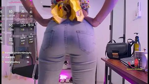 Media: Video of a woman with a yellow bow in her hair, wearing tight light blue jeans, standing in a modern room with a white wall and wooden table.