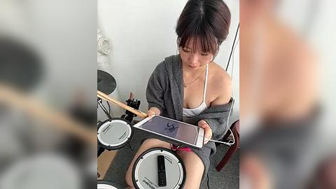 Media: A video of a young Asian woman with medium-length dark hair, wearing a gray robe and a white top, playing a digital drum set in a cluttered room.