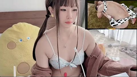 Media: A video of an Asian woman with long brown hair, wearing a cow-patterned bra and brown cardigan, sitting on a bed with a yellow stuffed animal.