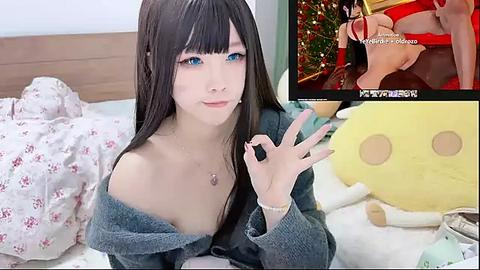 Media: Video of an Asian woman with long black hair, blue eyes, and fair skin, wearing a blue off-shoulder sweater, making an \"OK\" hand gesture. Background includes a bed with a white comforter and plush toys, including a large yellow bear.