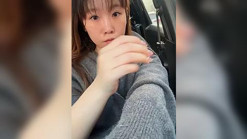 Media: Video of an East Asian woman with light skin, straight brown hair, and bangs, wearing a gray sweater, sitting in a car, holding her elbow, looking concerned.