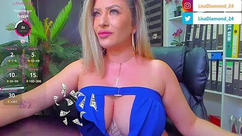 Media: Video of a blonde woman with large breasts wearing a revealing blue dress, sitting in an office with a potted plant, bookshelves, and a computer screen displaying a live feed.