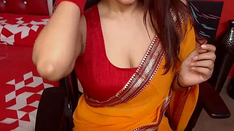 Media: Video of a light-skinned woman in a bright red sleeveless blouse and a yellow sari with gold embroidery, sitting on a red couch with white geometric-patterned cushions, holding a phone.