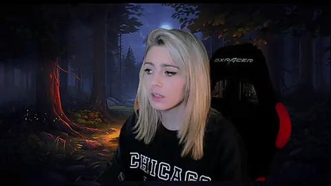 Media: A video of a young, blonde woman with straight hair, wearing a black \"Dignitas\" hoodie, sitting in a dark, dimly lit forest with a glowing tree to her left.