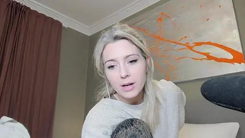 Media: Video of a young Caucasian woman with blonde hair, wearing a gray sweater, standing in a modern bedroom with abstract art and brown curtains.