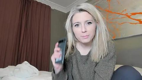 Media: A video of a blonde woman with fair skin, wearing a gray sweater, holding a smartphone. She sits on a bed with white sheets and a brown curtain in the background.